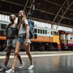 Two women with backpacks walking and smiling at a railway station.